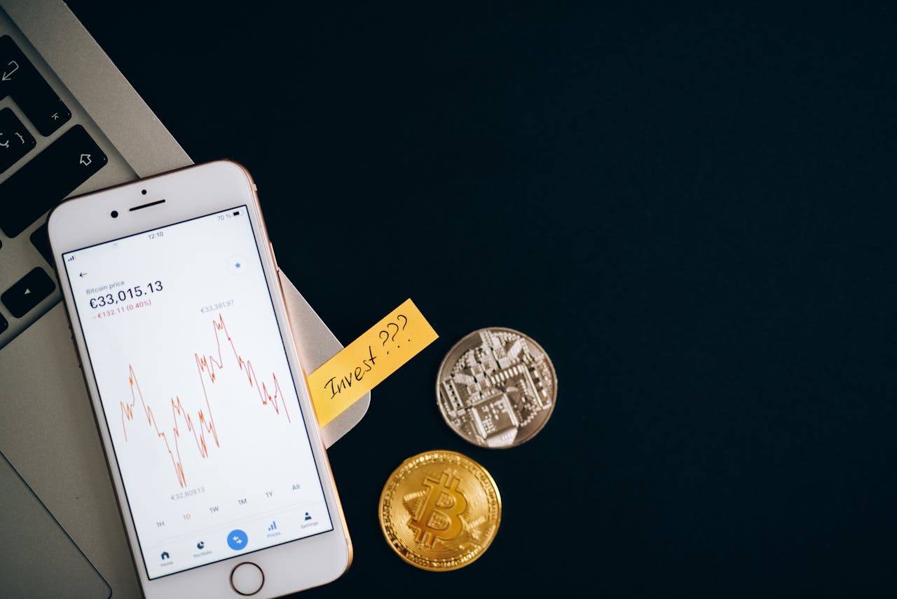 A smartphone displaying cryptocurrency prices beside coins and a laptop, symbolizing investment.
